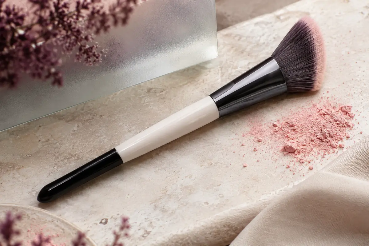Pinceau à blush biseauté noir et blanc aux poils denses, posé sur une pierre claire avec un pigment rose.