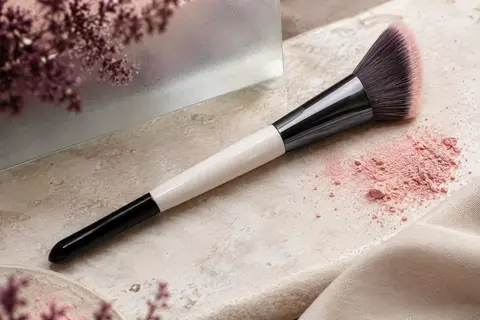 Pinceau à blush biseauté noir et blanc aux poils denses, posé sur une pierre claire avec un pigment rose.