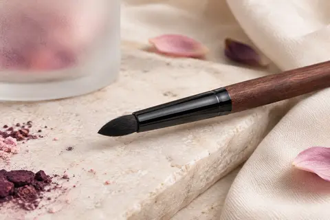 Pinceau eyeliner noir à tête dense et pointue posé sur une pierre claire près d’un verre dépoli.