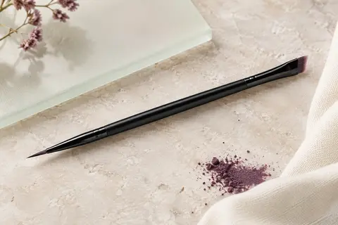 Pinceau de maquillage noir à double embout fin et biseauté posé sur une pierre claire