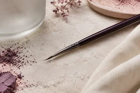 Pinceau eyeliner noir à tête faucille fine posé sur une surface en pierre claire.