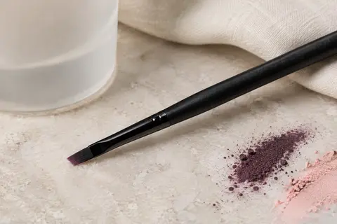 Pinceau eyeliner Eigshow à manche noir et tête fine inclinée posé sur une pierre claire.