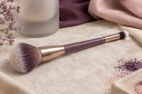 Pinceau IT Cosmetics à double embout posé sur une pierre claire, avec ses têtes synthétiques large et précise visibles.