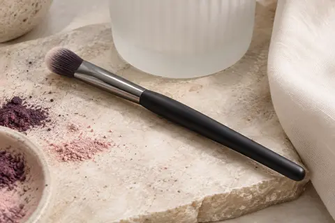 Pinceau pour les yeux à pointe arrondie, manche noir mat et virole gunmetal posé sur une pierre claire.