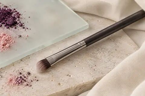 Pinceau plat pour paupières à fibres synthétiques posé sur une pierre claire près de pigments poudrés.