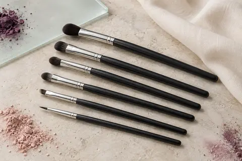 Six pinceaux noirs pour les yeux disposés sur une pierre claire avec quelques pigments prune et rose