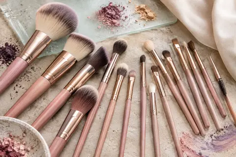 Assortiment de pinceaux de maquillage synthétiques à manches en bois rose doré, posé sur une pierre claire.