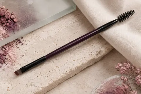 Pinceau à sourcils double-embout noir, tête biseautée et goupillon posés sur une pierre claire.