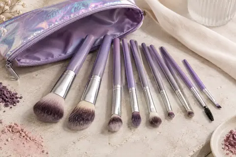 Dix mini-pinceaux de maquillage violets et leur trousse zippée irisée posés sur une pierre claire