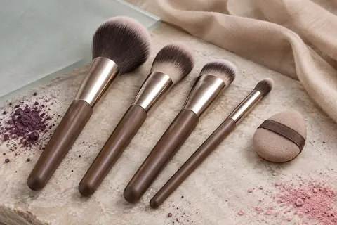 Quatre pinceaux de teint marron et une mini houppette disposés sur une surface en pierre claire