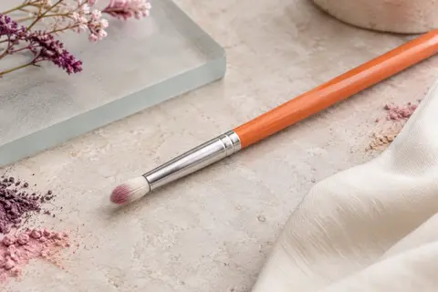 Pinceau correcteur à tête effilée, fibres synthétiques et manche orange posé sur une pierre claire