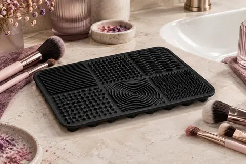 Tapis de nettoyage noir en silicone avec reliefs texturés, posé près de pinceaux sur une pierre claire.