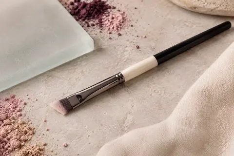 Petit pinceau correcteur plat biseauté aux fibres synthétiques, posé sur une pierre claire près de verre dépoli.