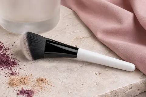 Pinceau blush biseauté noir et blanc posé sur une pierre claire près d’un tissu rose poudré.