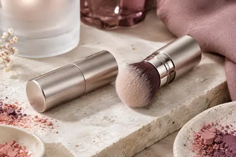 Pinceau kabuki rétractable à tête ronde posé sur une pierre claire avec du verre dépoli et un tissu mat.