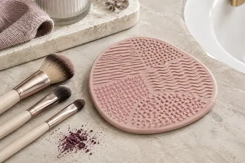 Tapis rose en silicone à reliefs texturés posé sur une pierre claire près de pinceaux de maquillage